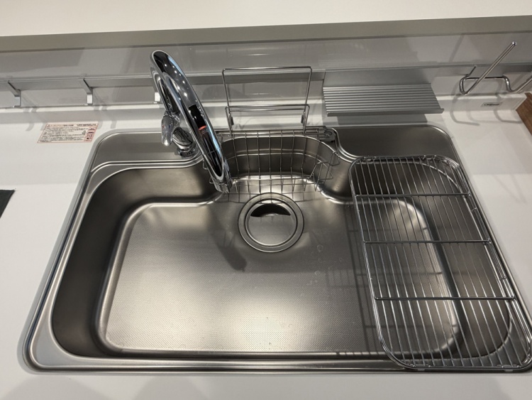 ステンレス製のシンクには、大きな蛇口と、食器を乾かすためのワイヤーラックが設置されています。清潔で使いやすそうなキッチンの一部です。