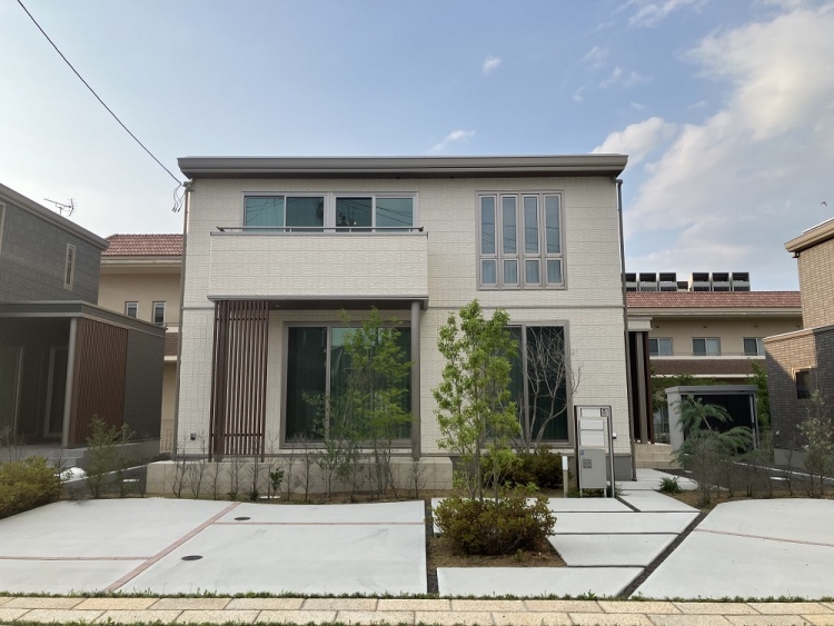 白い外壁の二階建て住宅の外観。前庭には植栽が植えられています。駐車場も見えます。