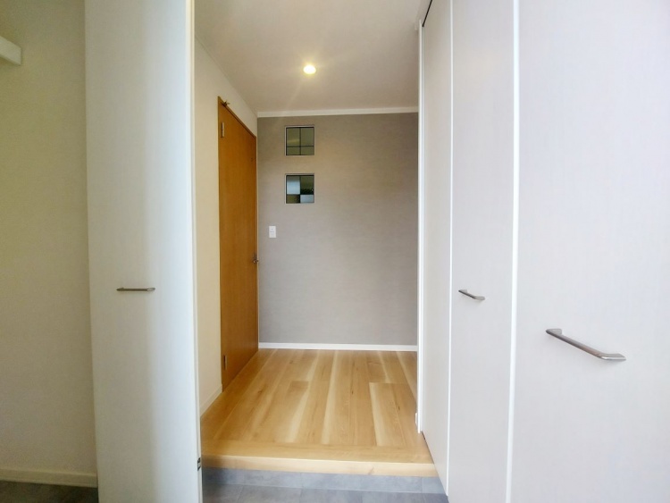 白い収納扉に囲まれた玄関スペース。木の床があたたかく迎えてくれ、奥の壁には四角い飾り窓が並んでいます。