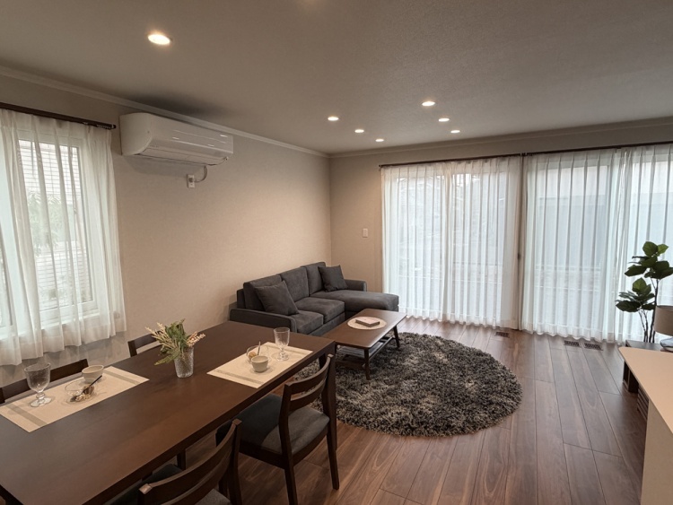 大きな窓から光が差し込むリビングダイニング。右側に灰色のソファがあり、その前には円形のラグとコーヒーテーブルがあります。手前には食卓があります。