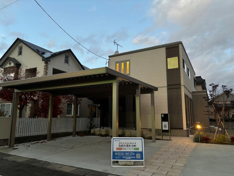 明るいベージュの外壁に黒い縦型のデザインパネルがアクセントとなっている二階建て住宅。前にはカーポートと広い駐車スペースがあり、植栽が家の周りを彩っています。青空の下、清潔感とスタイリッシュさが際立つ外観です。
