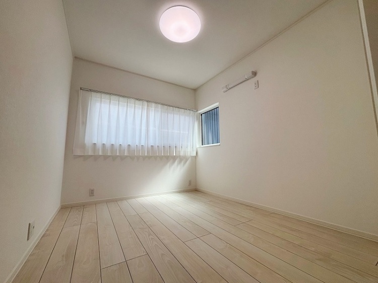 白い壁とカーテンが特徴のシンプルな洋室で、窓から柔らかい光が差し込んでいます。床は明るい色合いのフローリングで、落ち着いた雰囲気を感じさせます。