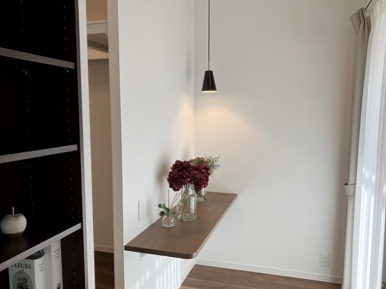 白い壁に木の棚が取り付けられ、ガラスの花瓶に赤い花が飾られた温かな一角です。