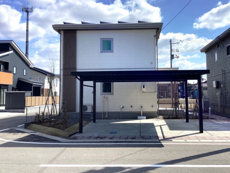 淡いグレーの外壁に木目のアクセントが映える2階建ての家が、角地に建っています。家の前にはカーポートがあり、植栽が施された庭が周囲を彩っています。