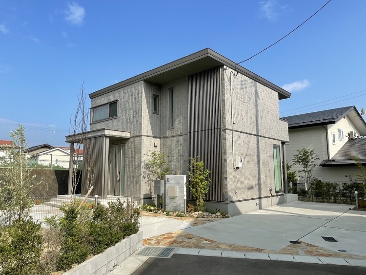 グレーの外壁が特徴的な2階建て住宅の外観。玄関前には縦格子の目隠しがあり、階段を上がった先に玄関があります。庭には低木や石が配置され、外観はすっきりとした印象です。