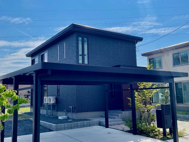 ダークグレーを基調とした外壁を持つ2階建て住宅の外観。前にはカーポートや緑豊かな庭が前景に広がっています。空には青空が広がり、静かな住宅街の一角が描かれています。