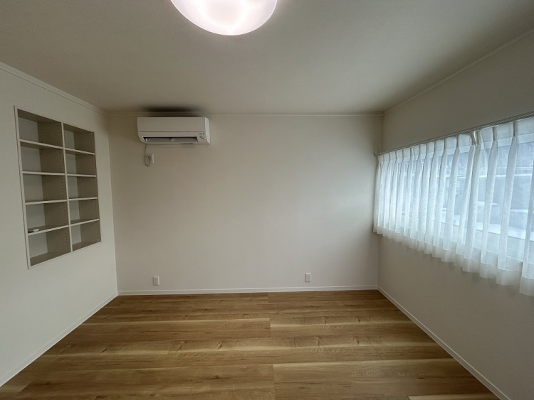 広々とした明るい部屋。壁一面の大きな窓から柔らかな光が差し込み、白いカーテンがふんわりと揺れる。木目の美しい床と壁の収納棚が、シンプルながらも温かみのある空間を作り出している。