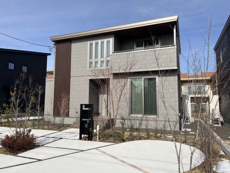 グレーの外壁にダークな木目調格子を基調とした２階建て住宅の外観。玄関の前には小庭、植栽が植えられています。手前には駐車場が見えます。