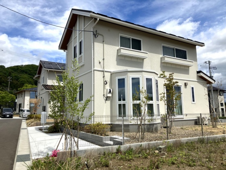 白い外壁の2階建て住宅が晴れ空の下に建っています。小さな前庭と駐車スペースがあり、玄関には黒い縦格子がアクセントとなっています。住宅は静かな住宅地に位置しています。