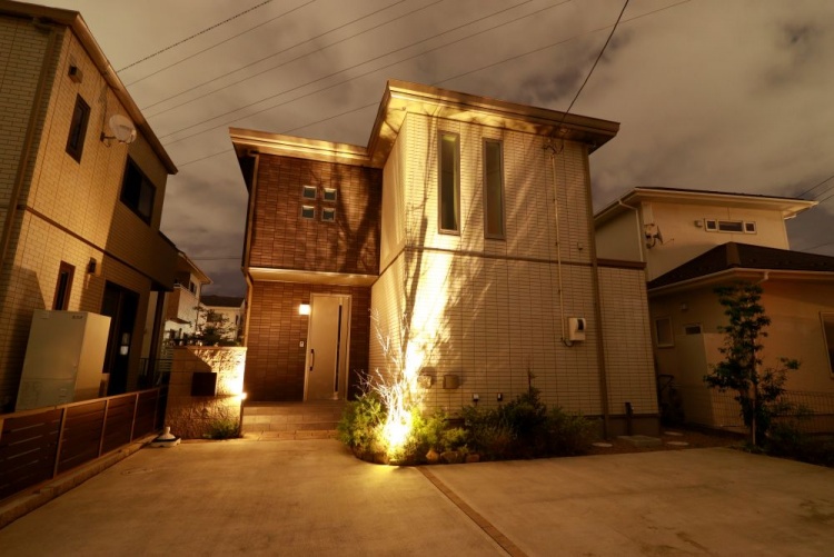 あたたかな光が玄関まわりを包み込む夜の住宅。空には雲が広がり、植栽のライトアップが幻想的な陰影を描いています。