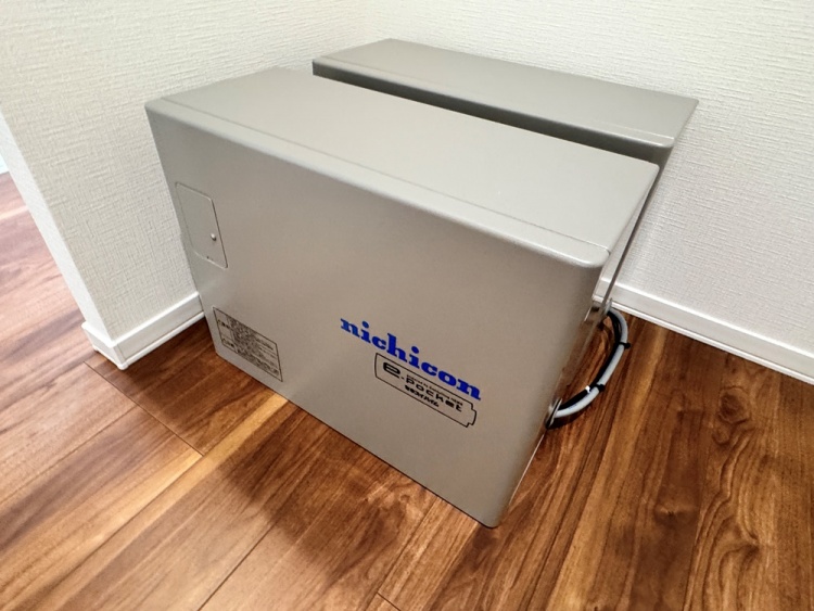 床に設置されたグレーの蓄電池で、壁に沿って設置されています。機器には「nichicon」と青い文字が書かれており、コードが壁に繋がっています。