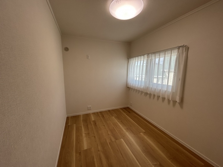 ナチュラルな木目の床が広がる、落ち着いた雰囲気の部屋。壁は優しい色合いで統一され、大きな窓から外の景色が見える。レースカーテン越しに柔らかな光が差し込み、穏やかな空間を演出している。