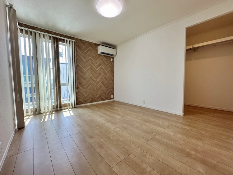 木目調のアクセント壁と縦型ブラインドが印象的な部屋。床はナチュラルな色合いのフローリングで、エアコンが設置されています。奥にはウォークインクローゼットがあります。