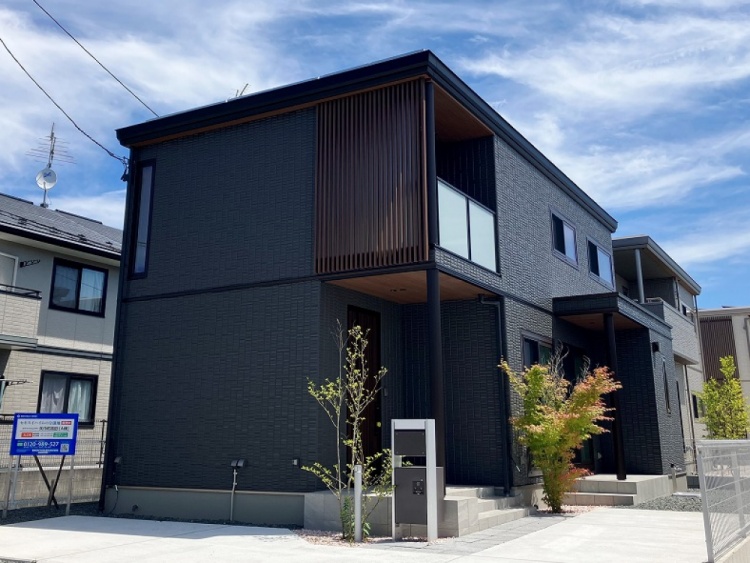 濃いグレーの外壁が特徴的な2階建住宅の外観。縦長の窓と木製の格子がアクセントになっています。前庭には若い木々が植えられており、清潔なコンクリートの駐車スペースがあります。