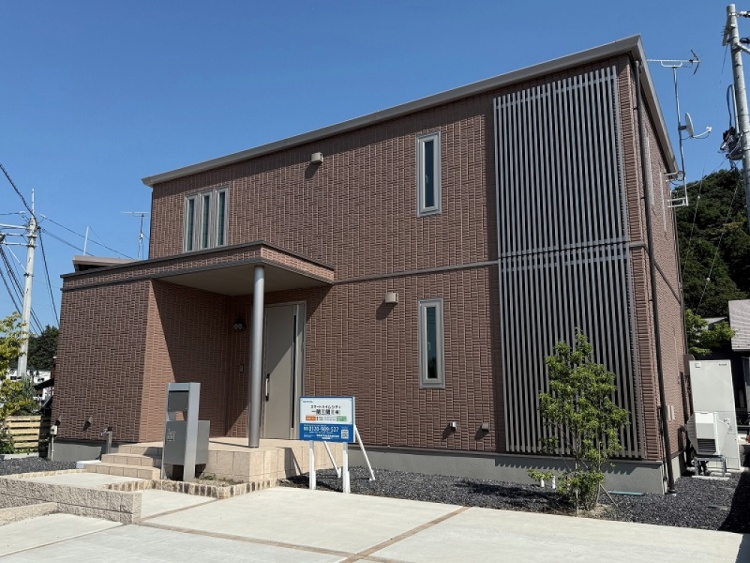 ブラウンを基調とした二階建て住宅の外観。玄関には庇があり、玄関前には階段が設けられています。縦長の窓と、右側のルーバー状の外壁デザインが特徴です。敷地内には植栽が点在しています。