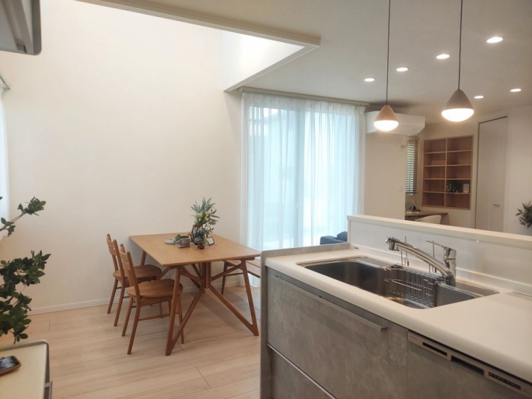 ナチュラルな木目調のフローリングの上に木製のダイニングテーブルと椅子が置かれたダイニングエリア。奥にはブルーグレーのソファを置いたリビングがあり、大きな窓から光が差し込む明るい空間。天井にはダウンライトが設置されている。