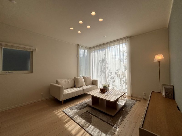 広い窓のある明るいリビングに、白いソファとセンターテーブル、スタンドランプが配置されています。