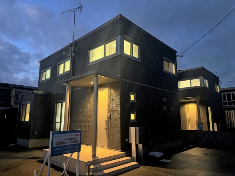ダークグレーを基調とした二階建て住宅の外観で、シンプルなデザインが特徴です。玄関には小さな階段があり、縦長の窓がスタイリッシュに配置されています。