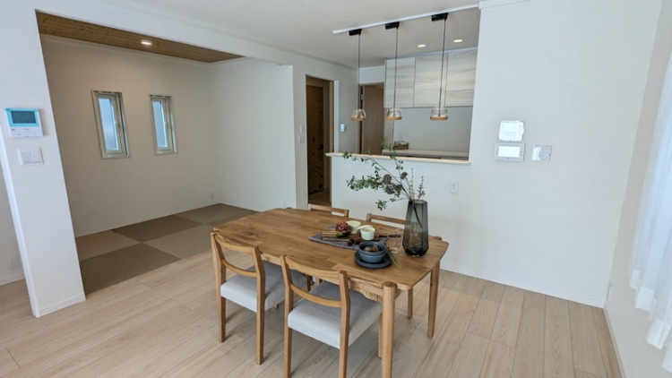 木目調を基調としたダイニングキッチン。ダイニングテーブル、椅子が配置されています。窓、収納が設置されています。左側に畳スペース、奥にキッチンが見えます。