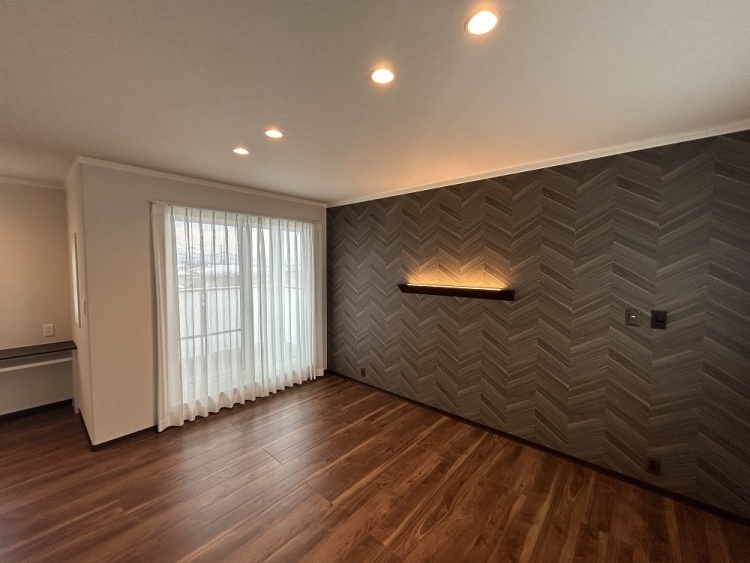 広々とした部屋に、大きな窓から柔らかな光が差し込む。ヘリンボーン柄のアクセントウォールが上品な雰囲気を演出し、間接照明が優しく照らしている。床は温かみのある木目調。