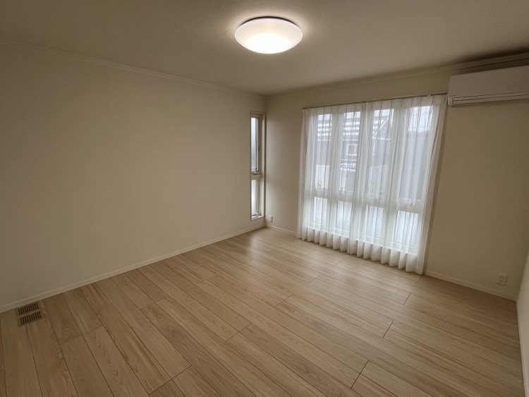 広々とした部屋に、大きな窓から自然光が差し込む穏やかな空間。白いカーテンが優しく揺れ、木目の床が温もりを感じさせる。壁にはエアコンが設置され、快適な居住空間が広がっている。