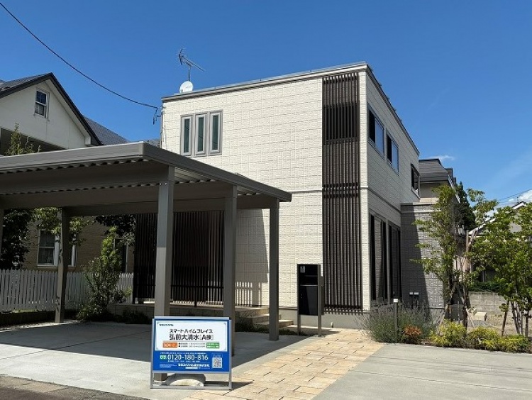 明るいベージュの外壁に黒い縦型のデザインパネルがアクセントとなっている二階建て住宅。前にはカーポートと広い駐車スペースがあり、植栽が家の周りを彩っています。青空の下、清潔感とスタイリッシュさが際立つ外観です。