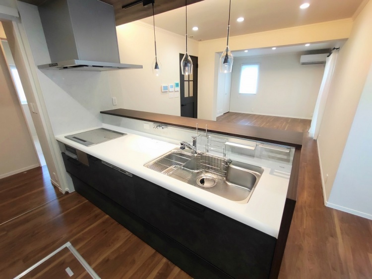 広々としたキッチン空間。ダークカラーの収納と白いカウンターが調和し、モダンな雰囲気を演出。奥には大きな窓があり、柔らかなカーテン越しに自然光が広がる。