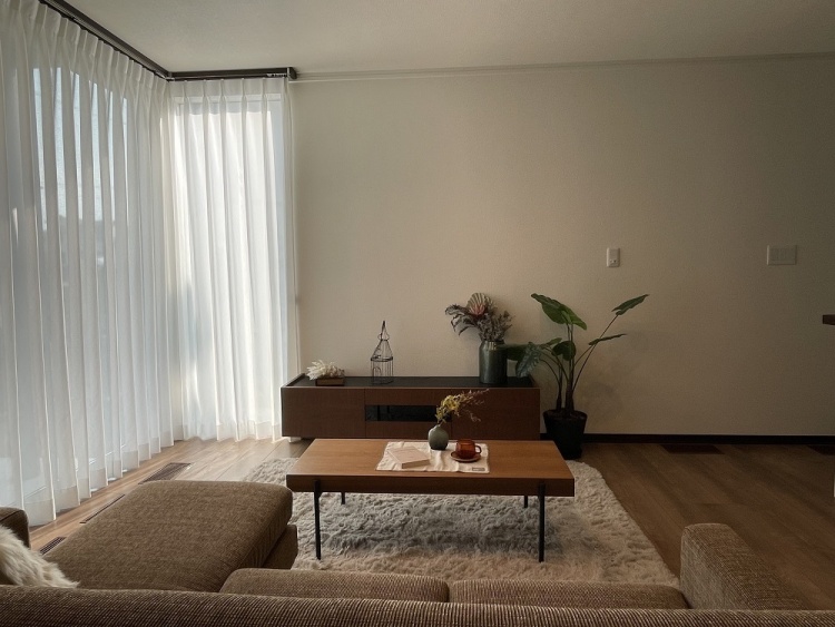 明るい窓際のリビングルームで、落ち着いた木製家具と観葉植物が置かれています。テーブルの上には小物が並び、窓からの光が部屋全体に広がり、リラックスできる雰囲気を演出しています。