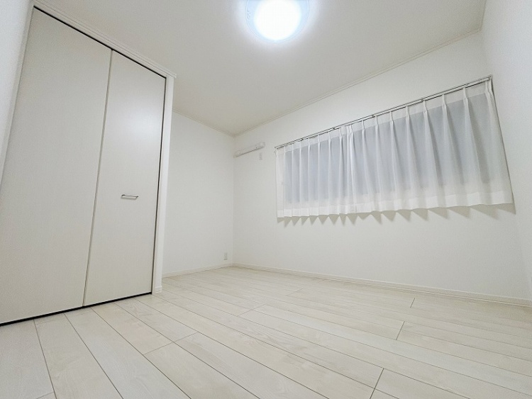 白い壁とカーテンが特徴のシンプルな洋室で、窓から柔らかい光が差し込んでいます。床は明るい色合いのフローリングで、落ち着いた雰囲気を感じさせます。