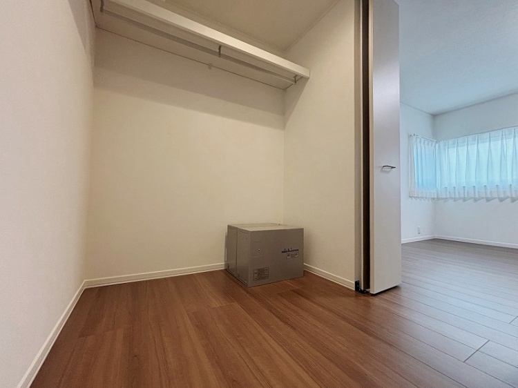 広々としたウォークインクローゼット。白い棚とハンガーパイプが備え付けられ、衣類をたっぷり収納できる。床は落ち着いた木目調で、部屋全体が明るく清潔な印象。隣の部屋には窓があり、柔らかな光が差し込んでいる。