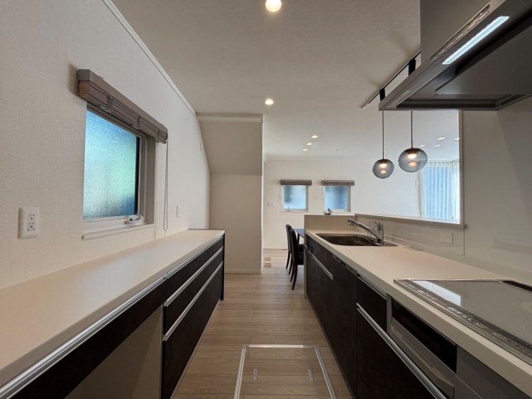 ダークな木目調を基調としたキッチン。シンクの背面には家電カウンターと食器棚が配置されています。ペンダイトライト、小窓が見えます。奥にはダイニングが見えます。