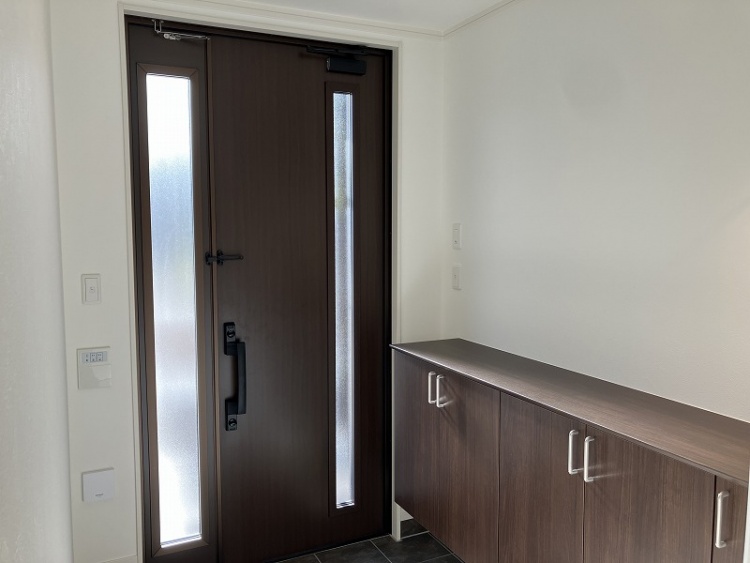 広々とした玄関には、ダークブラウンのキャビネットが設置され、ペンダントライトが壁に温かい光を照らしています。白いタイルの床が清潔感を演出しています。