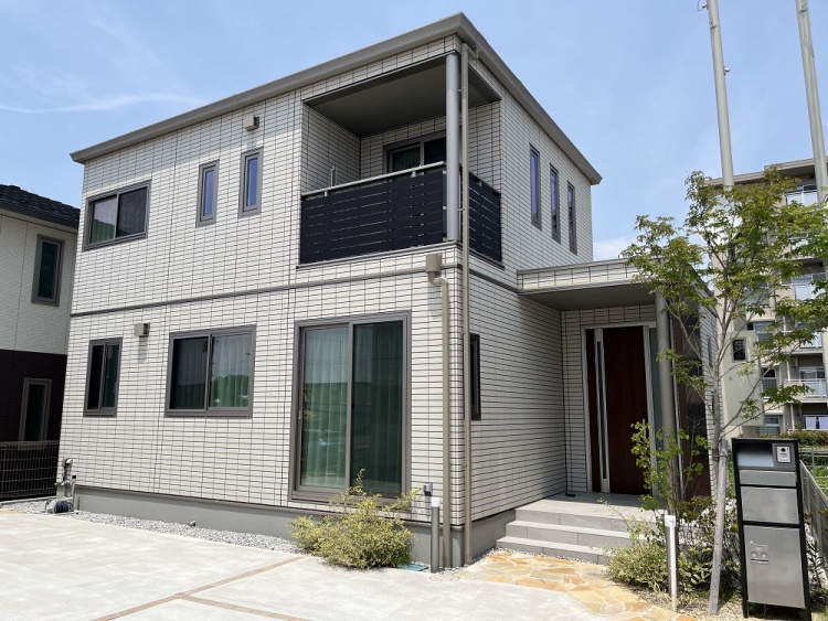 白い外壁の２階建て住宅の外観。大きな窓とバルコニーがあり、庭には植栽とポストが配置されています。植栽が植えられています。