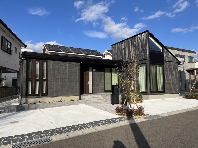 青空の下に建つ平屋の住宅。黒い外壁と大きな窓が特徴的で、片流れの屋根には太陽光パネルが設置されている。玄関周りには植栽が配置され、落ち着いた雰囲気を演出している。
