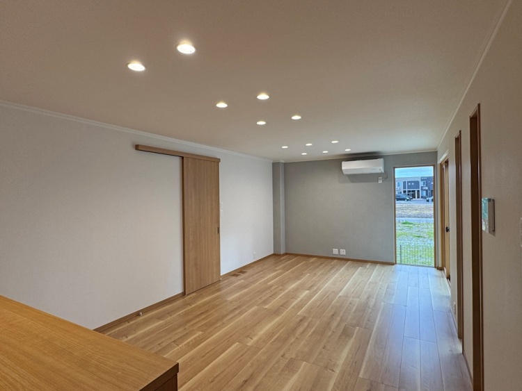 温かみのある木目の床が広がるシンプルなリビング。天井には複数のダウンライトが並び、均一な明るさを提供。奥には外へつながるドアがあり、開放感を感じる。左側には引き戸があり、別の空間へと続いている。