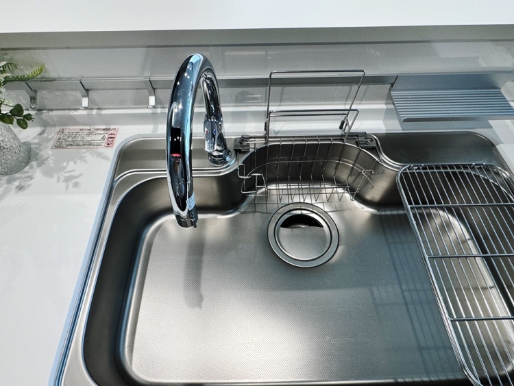 ステンレス製のシンクには、大きな蛇口と、食器を乾かすためのワイヤーラックが設置されています。清潔で使いやすそうなキッチンの一部です。