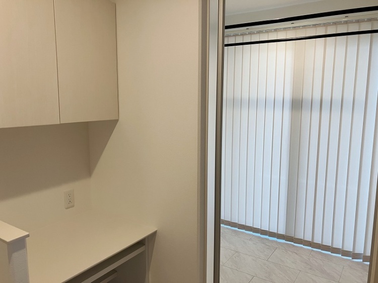 白を基調としたすっきりとした収納スペース。壁に取り付けられた収納棚とカウンターがあり、作業や家電の設置に便利。床は大理石調のタイルが敷かれ、清潔感のある印象。奥には大きな窓があり、バーチカルブラインドが柔らかな光を演出しています。