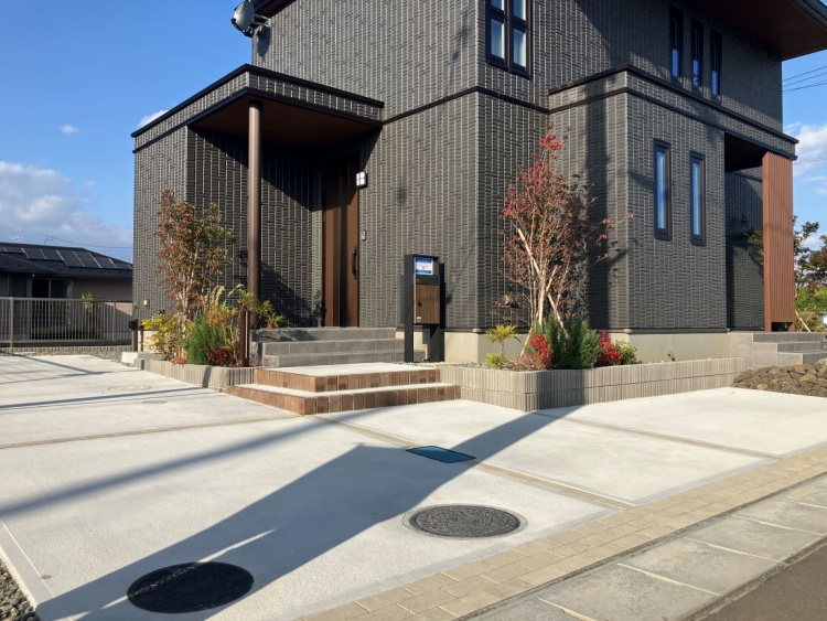 広い駐車スペースと段差のある玄関がある家の外観で、植えられた若木がやさしく風に揺れています。