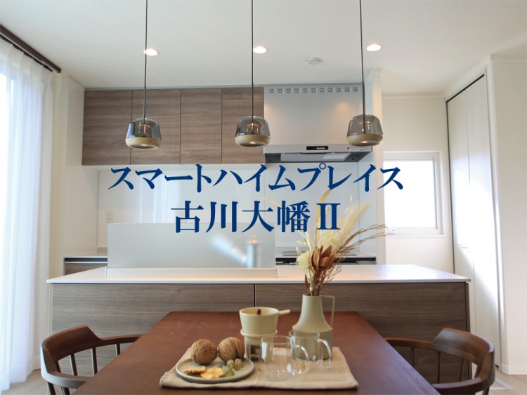 心地よい光が差し込むキッチンとダイニング。落ち着いた木目調の収納と、モダンなペンダントライトが洗練された印象を与える。テーブルには温かみのある食器やドライフラワーが並び、くつろぎの時間を演出。背景には「スマートハイムプレイス 古川大幡Ⅱ」の文字が記されている。