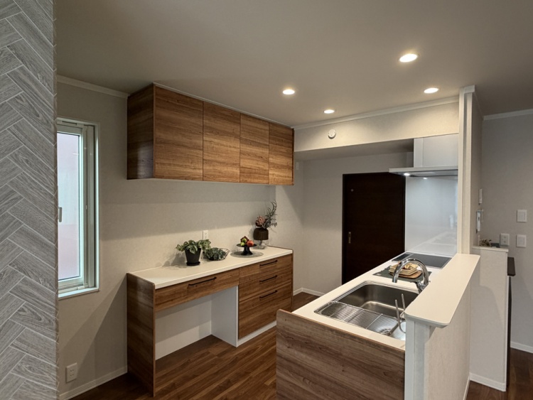 ダークな木目調を基調としたキッチン。シンクの背面には家電カウンターと食器棚が配置されています。中央右には洗面脱衣室が見えます。