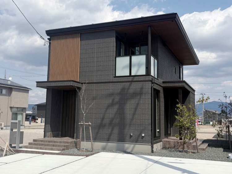 ダークグレーの外壁を持つ2階建ての住宅が、夕暮れの下に建っています。前には小さな庭があり、若木が植えられています。