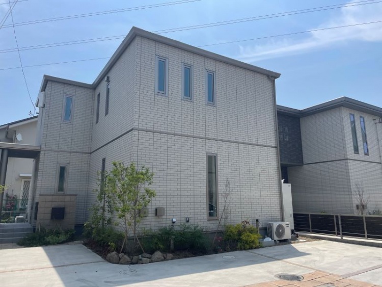 白い外壁の二階建て住宅の外観。前には植栽とエアコンの室外機が見えます。