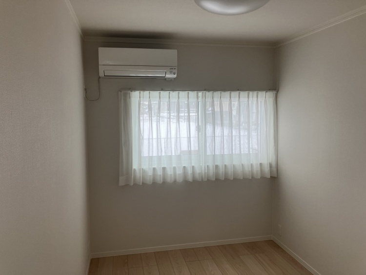 コンパクトな部屋に木目調の床が広がり、壁にはエアコンが設置されています。小さめの窓には白いカーテンがかかり、優しい光が差し込んでいます。落ち着いた雰囲気の空間です。