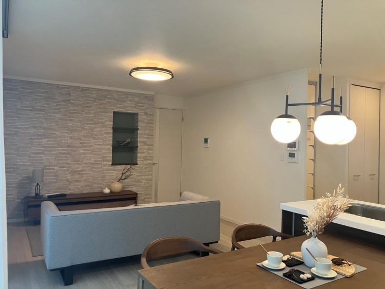 ペンダントライトが印象的な木製ダイニングテーブルと、奥にグレーのソファが配置されたナチュラルモダンなLDK。キッチンは対面式で、白い天板とダークブラウンの収納が特徴的。石目調のアクセントウォールがあるリビングには間接照明もある。