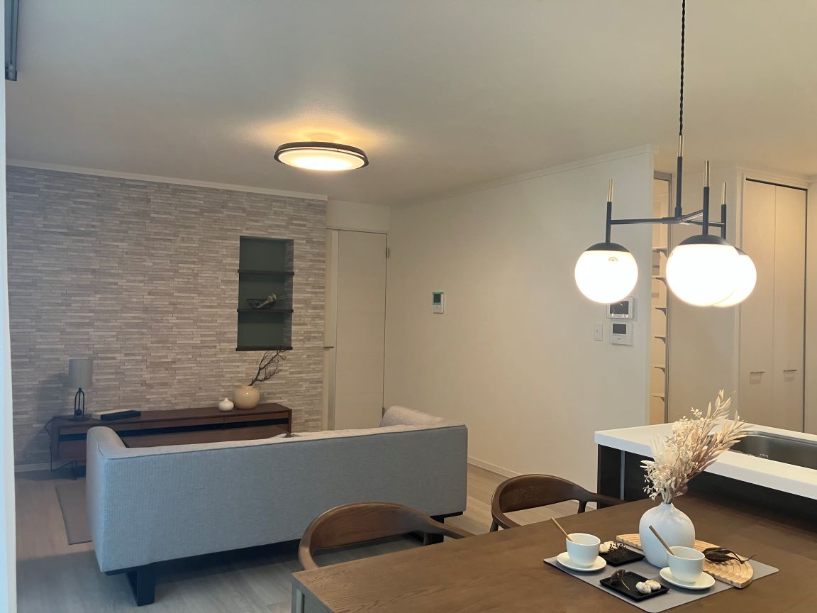 ペンダントライトが印象的な木製ダイニングテーブルと、奥にグレーのソファが配置されたナチュラルモダンなLDK。キッチンは対面式で、白い天板とダークブラウンの収納が特徴的。石目調のアクセントウォールがあるリビングには間接照明もある。