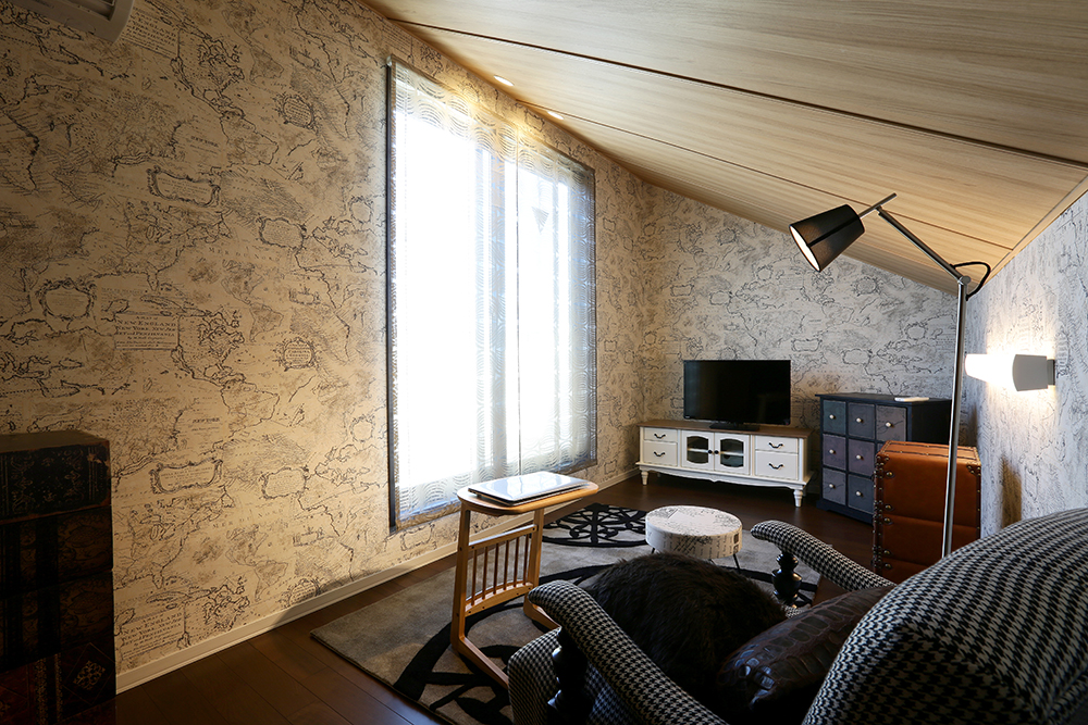 斜め天井の落ち着いた小屋裏スペース（コンファティック）。壁紙にはヴィンテージの地図模様が施され、アンティーク調の家具やランプが配置されています。小さなテレビと収納棚が置かれ、シックでリラックスした空間が広がっています。