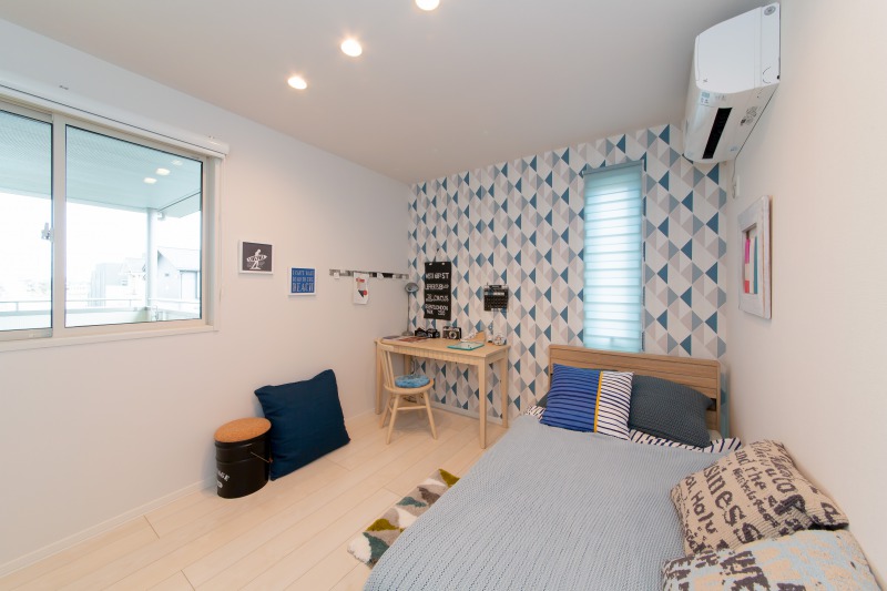 明るくポップなデザインの子供部屋です。壁には青と白の幾何学模様のアクセントがあり、ベッドとデスクがシンプルに配置されています。窓からは外の景色が見え、清潔感あふれる空間です。