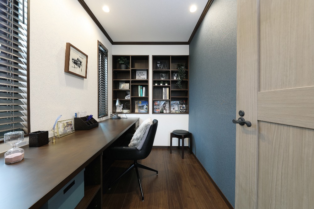 コンパクトな書斎の部屋に、壁際に長いデスクと黒い椅子が配置されています。奥の壁には本や飾りが並ぶ棚があり、左側の窓から自然光が差し込んでいます。シンプルで落ち着いた雰囲気のワークスペースです。