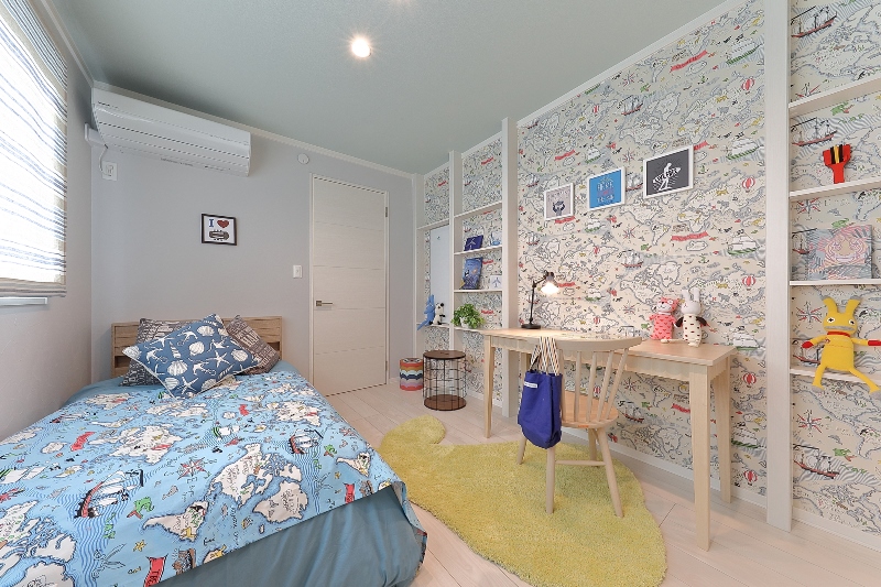 明るくかわいらしい子ども部屋。ベッドには世界地図が描かれた青い布団が掛けられ、壁にはカラフルな地図柄の壁紙が貼られています。木製のデスクと椅子があり、棚にはおもちゃや本が並んでいます。黄色いラグが床に敷かれ、楽しい雰囲気の部屋です。