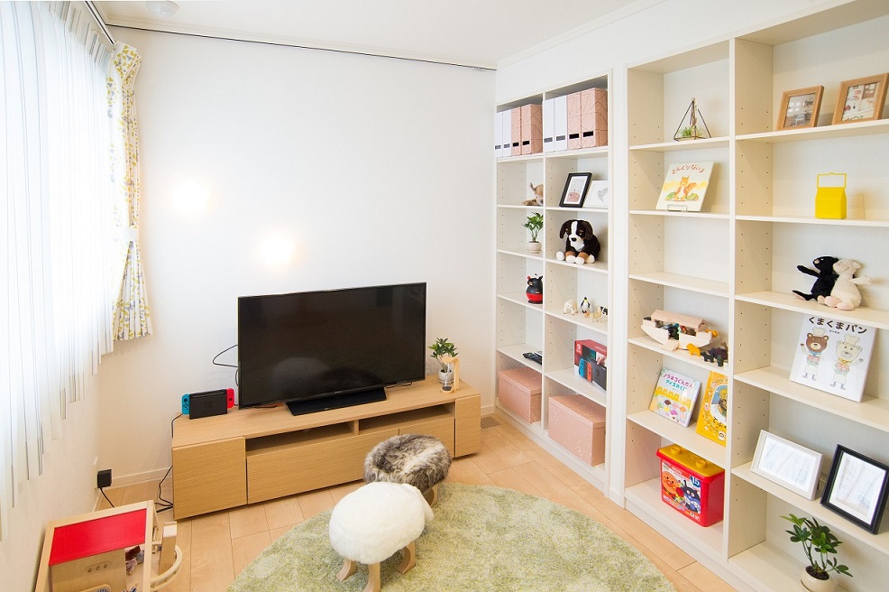 白を基調とした明るい子供部屋。テレビの横におもちゃや絵本が並ぶ棚があり、床には小さな可愛らしい椅子が置かれています。柔らかい光が部屋全体を照らしています。